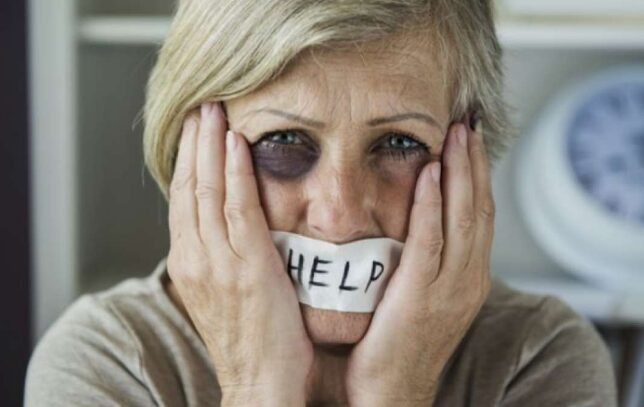 elderly woman with black eye holding a help sign on her lips